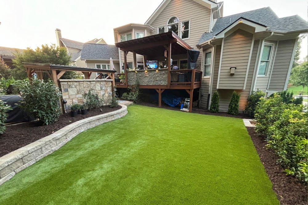 A backyard with an elevated wooden deck, stone accent wall, artificial turf lawn, and surrounding shrubs in front of a two-story house.