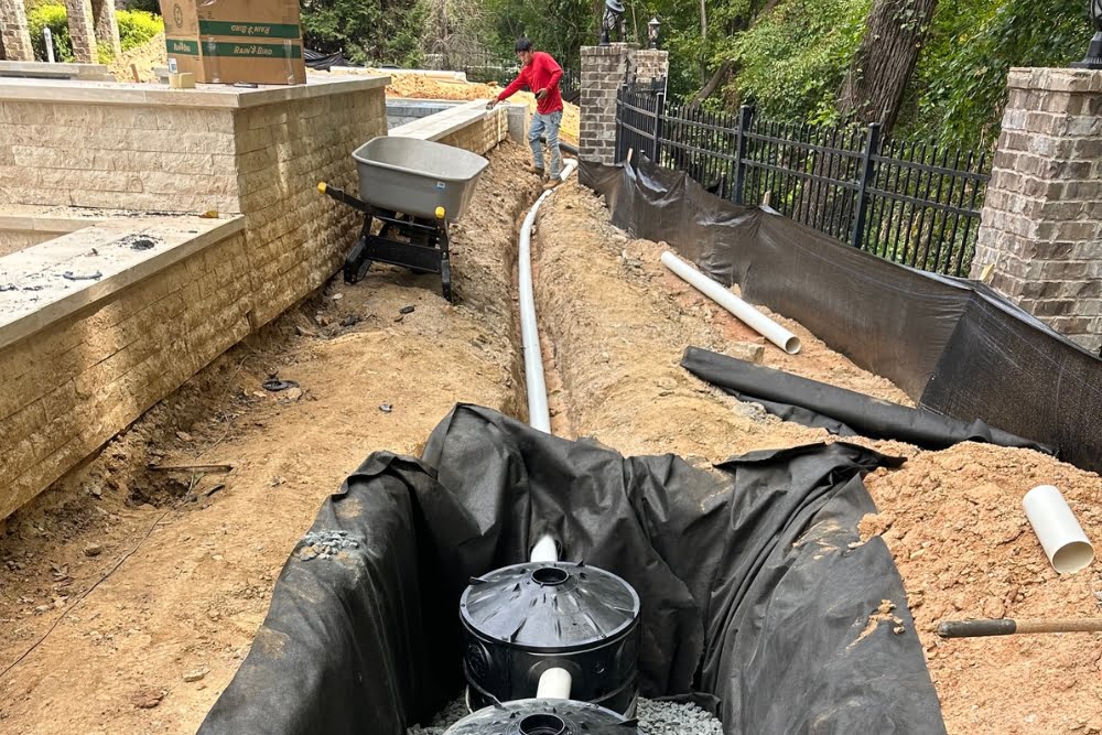 A person works on installing a drainage system, with pipes laid in a trench and a partially buried catch basin in the foreground. Construction materials and fencing are visible, showcasing the detailed landscaping services being executed.