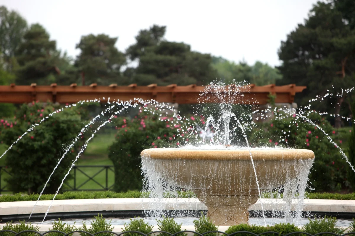 A stone fountain with multiple water jets surrounded by greenery and a wooden pergola in the background, all seamlessly blending into stunning outdoor entertainment spaces.