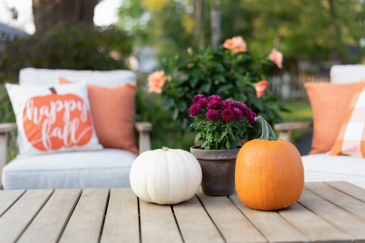 A wooden table displays a white and an orange pumpkin along with a pot of purple flowers. In the background, there are white chairs with orange cushions and a "happy fall" pillow, all set against the backdrop of thoughtfully crafted seasonal landscaping.