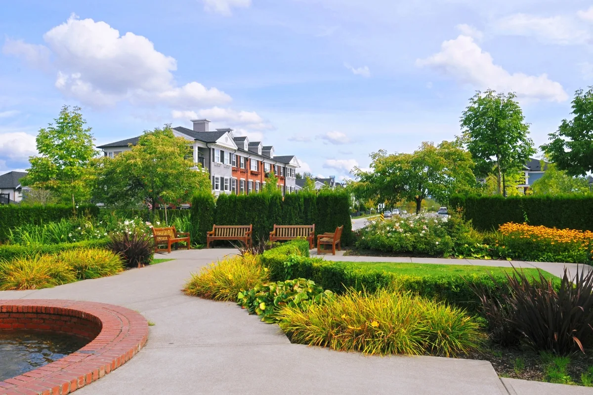 A landscaped garden featuring community green spaces, complete with benches, pathways, lush greenery, and blooming flowers. Residential buildings and a blue sky with fluffy clouds can be seen in the background.