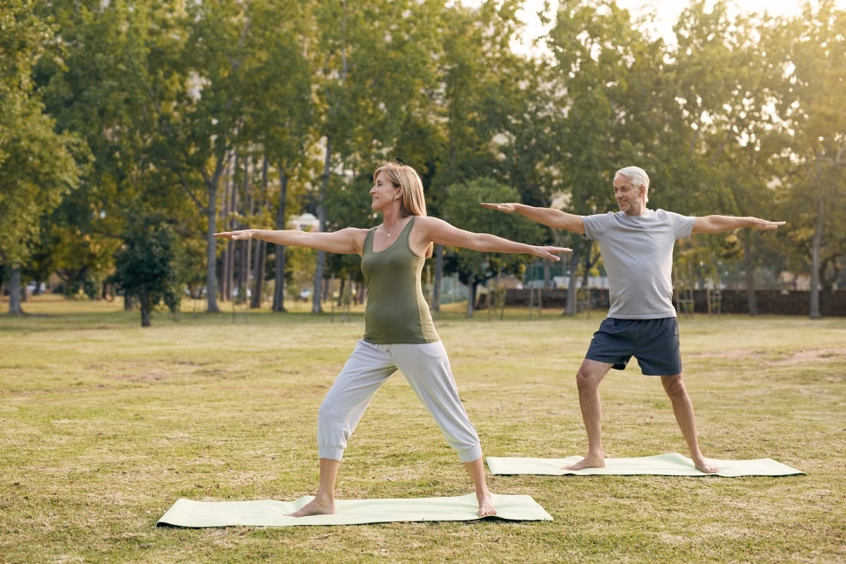 Two people practice yoga in the community green space, performing a standing pose with arms extended, surrounded by lush trees and vibrant grass.