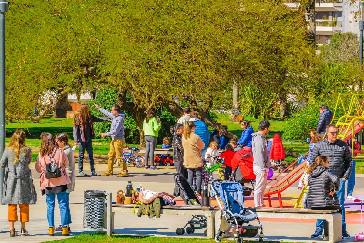 In community green spaces, people gather in a park with children playing on playground equipment, strollers nearby, and trees in the background.