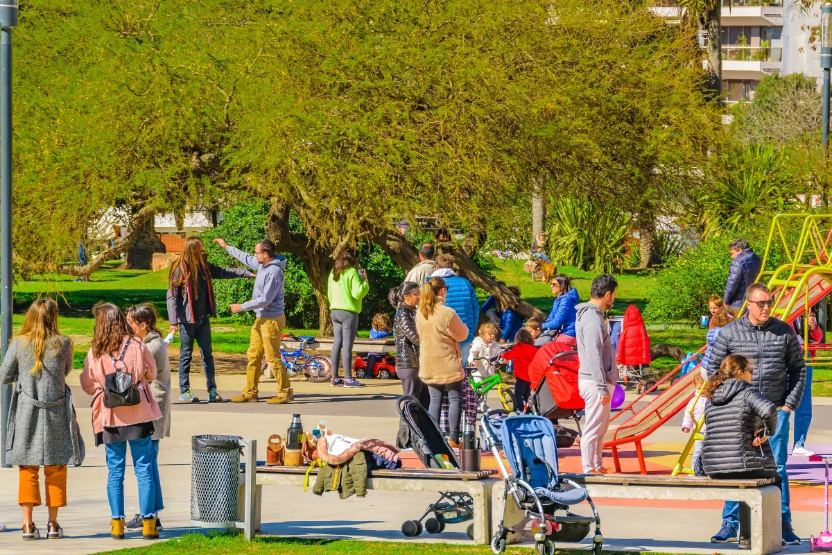 In community green spaces, people gather in a park with children playing on playground equipment, strollers nearby, and trees in the background.