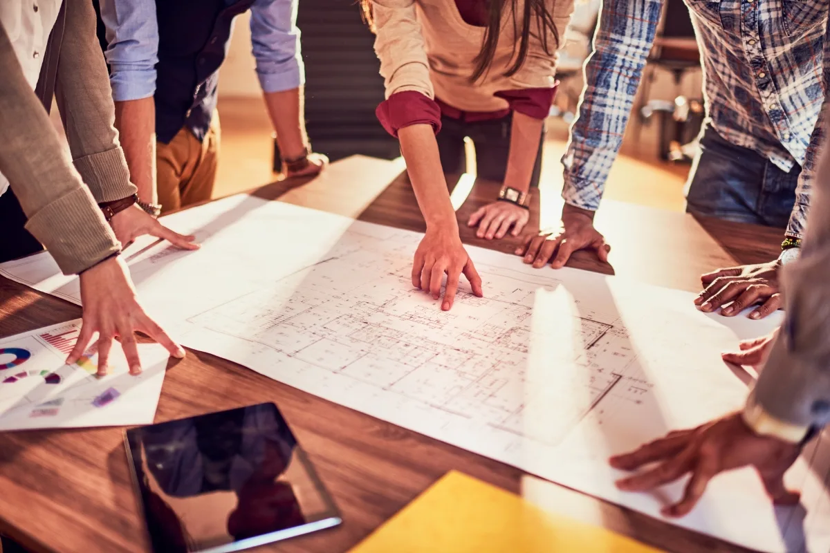 A group of people stands around a table reviewing architectural blueprints, envisioning future community green spaces, with a tablet and papers nearby.