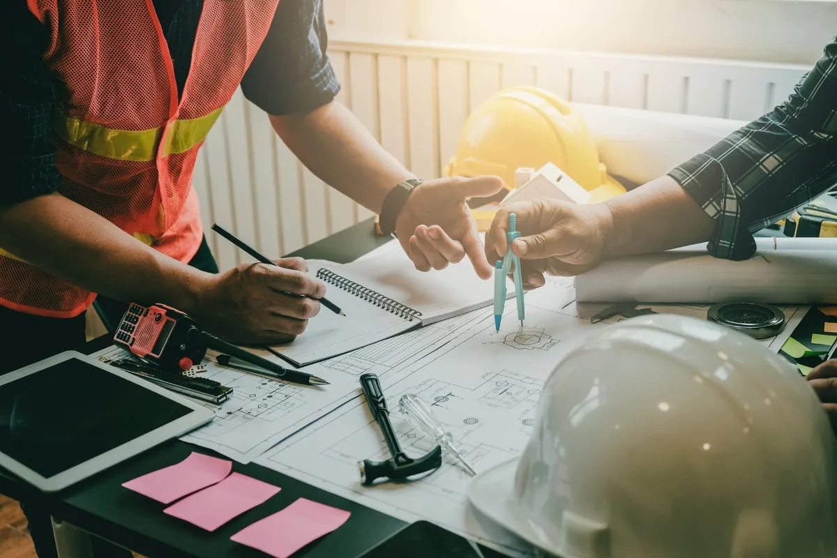 Two people in work attire discuss blueprints for community green spaces on a table with tools, a tablet, and safety helmets. One holds a compass, while the other takes notes.