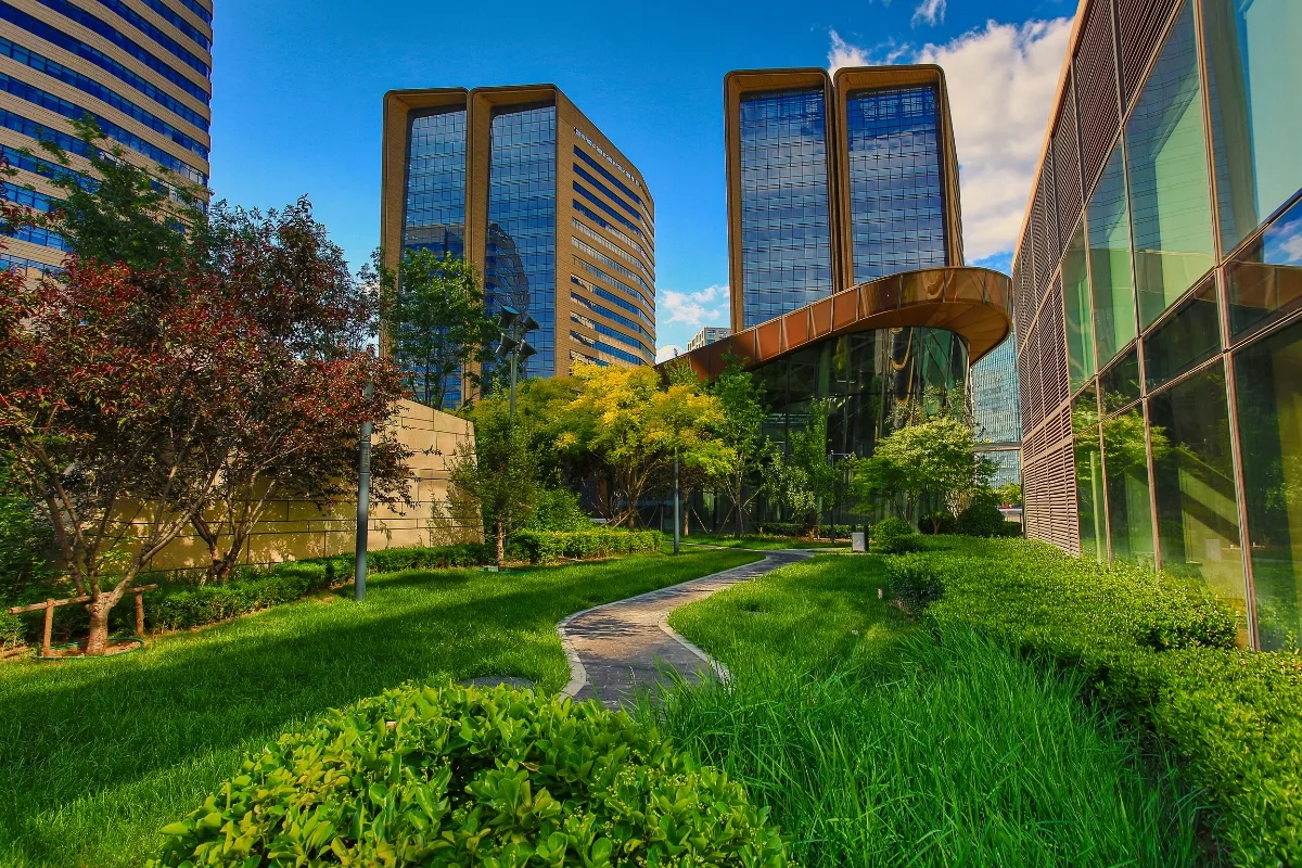 A modern office complex with sleek glass buildings is nestled amidst community green spaces, surrounded by lush lawns and trees under a clear blue sky.