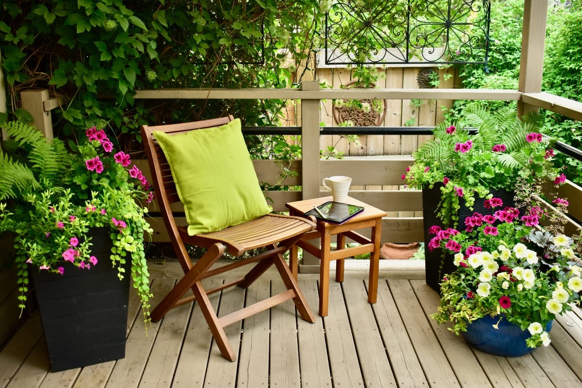 Wooden chair with a green cushion beside a small table holding a cup and tablet, set amidst lush plants and flowers on a wooden deck. Perfect for outdoor makeovers that transform your space into a serene retreat.