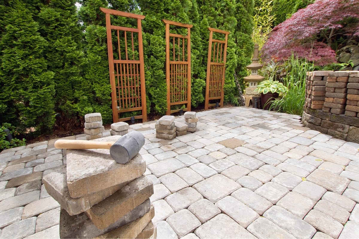 A stone-paved garden showcases a rubber mallet resting on stacked stones in the foreground, epitomizing outdoor makeovers. Wooden trellises and lush greenery beautifully frame this tranquil space.