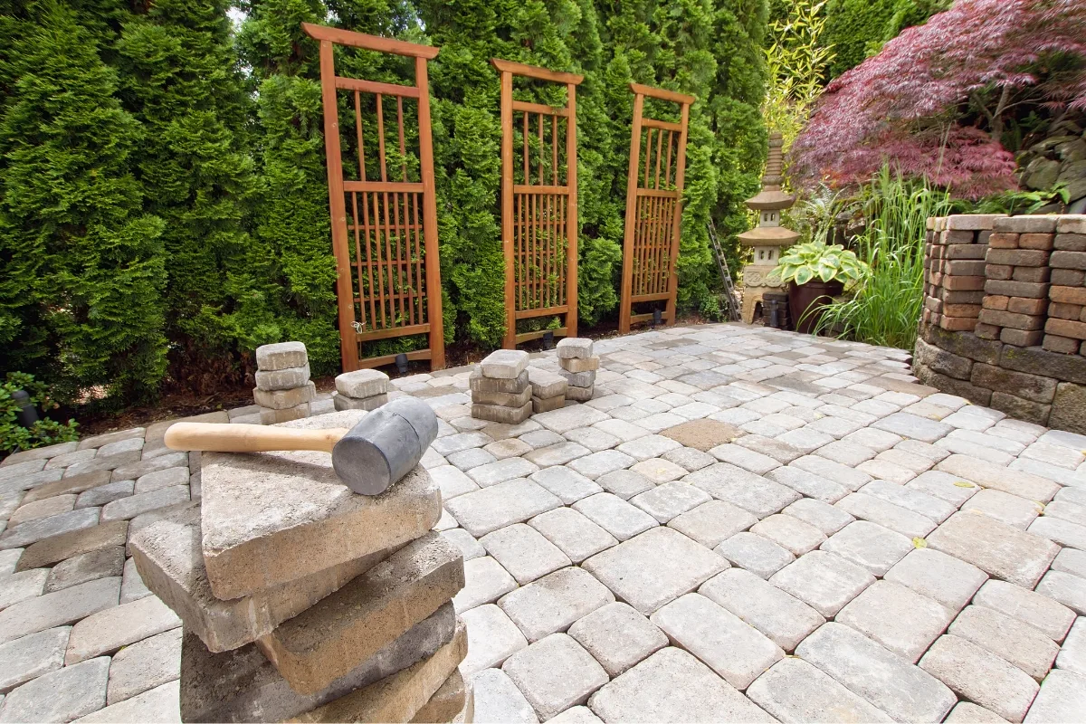 Outdoor Makeovers: Transform Your Yard with Elegant Landscaping & Hardscaping 3 A stone-paved garden showcases a rubber mallet resting on stacked stones in the foreground, epitomizing outdoor makeovers. Wooden trellises and lush greenery beautifully frame this tranquil space.
