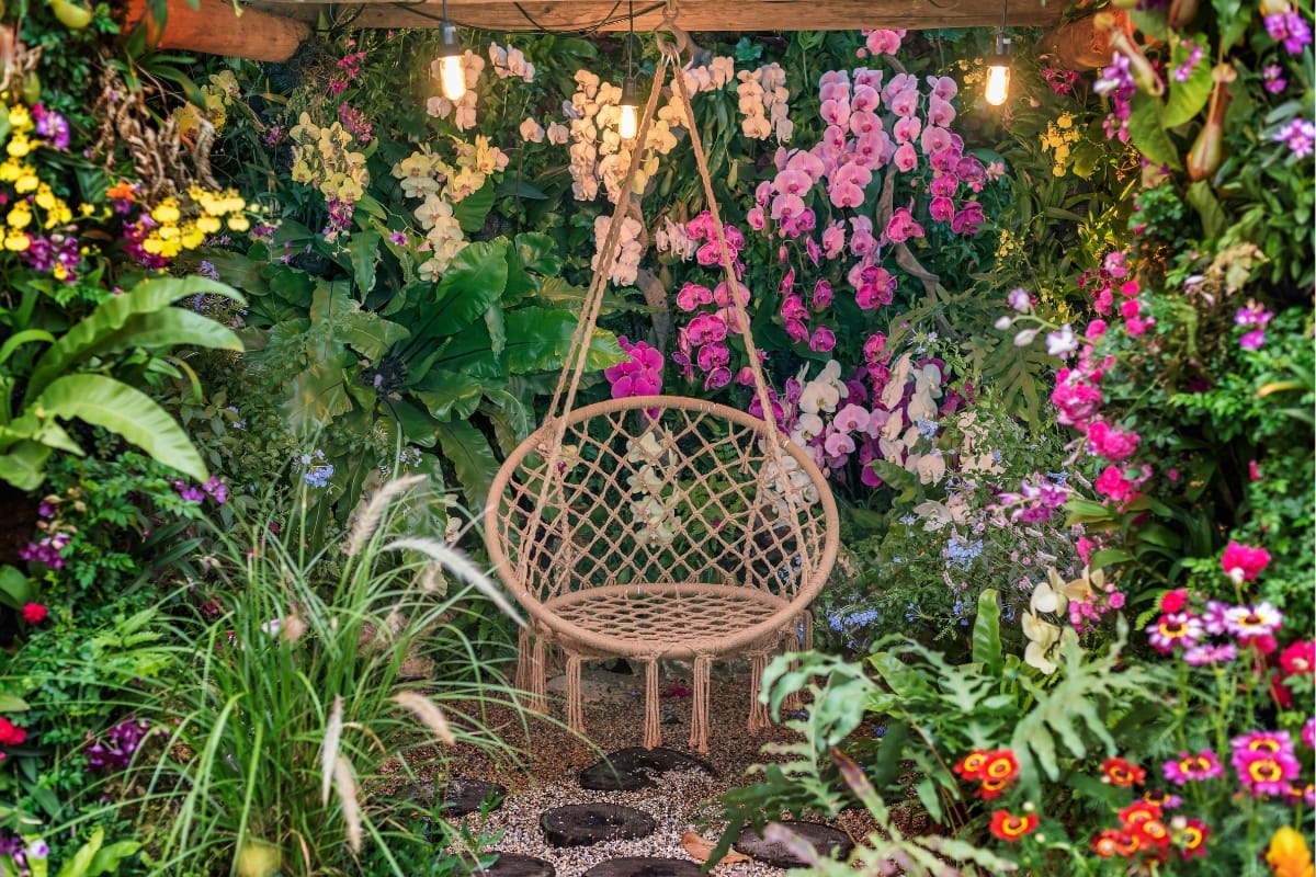 A wicker hanging chair graces the lush garden, a testament to outdoor makeovers, surrounded by vibrant flowers and foliage under twinkling hanging lights.