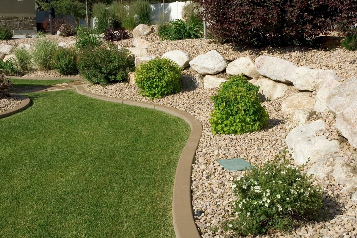 A landscaped garden with a curved grassy path bordered by stones, small bushes, and shrubs, set against a backdrop of larger rocks, is the epitome of outdoor makeovers.