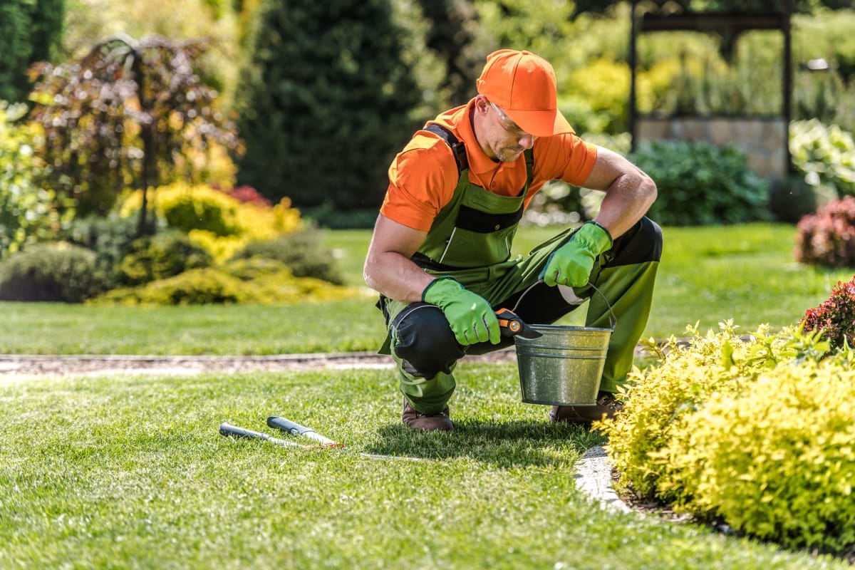 A gardener in an orange cap and uniform crouches with precision, working with tools near a bucket on a well-maintained lawn, surrounded by trees and shrubs—a scene of dedicated outdoor makeovers.