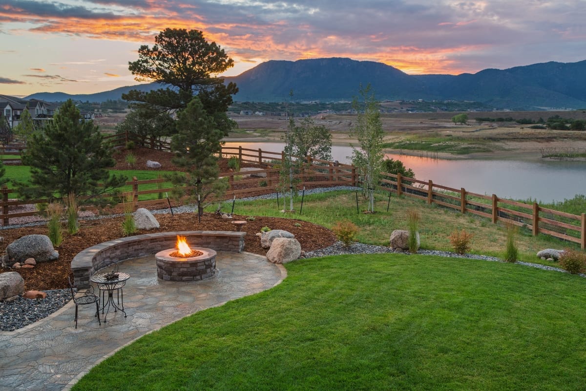 A landscaped backyard showcases a stunning outdoor makeover with a stone patio and lit fire pit, all surrounded by grass and rocks. A wooden fence frames the serene lake, set against mountains and a vibrant sunset backdrop.