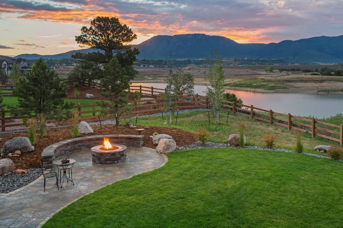 Outdoor Makeovers: Transform Your Yard with Elegant Landscaping & Hardscaping 1 A landscaped backyard showcases a stunning outdoor makeover with a stone patio and lit fire pit, all surrounded by grass and rocks. A wooden fence frames the serene lake, set against mountains and a vibrant sunset backdrop.