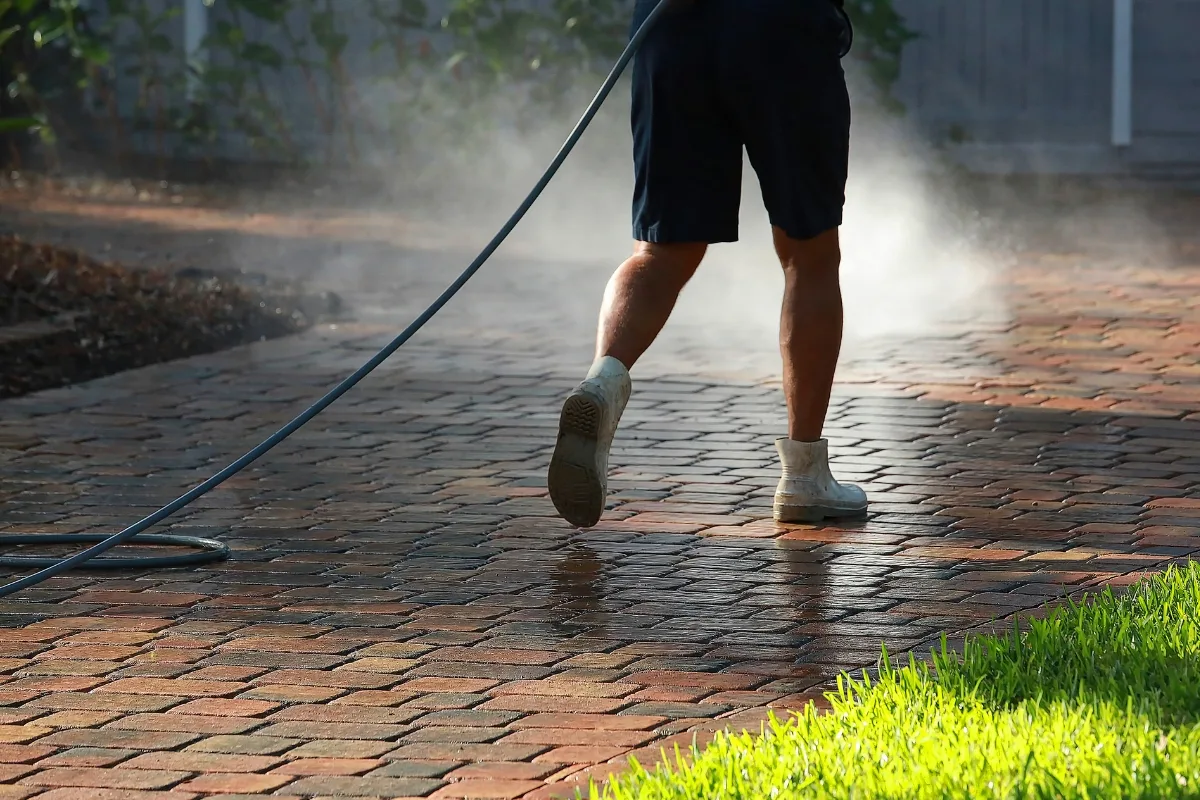 A person pressure washing a brick driveway, with water mist shimmering in the air. Dressed in shorts, socks, and sneakers, they seem to be preparing the surface meticulously&mdash;perhaps learning how to lay pavers for a new project.