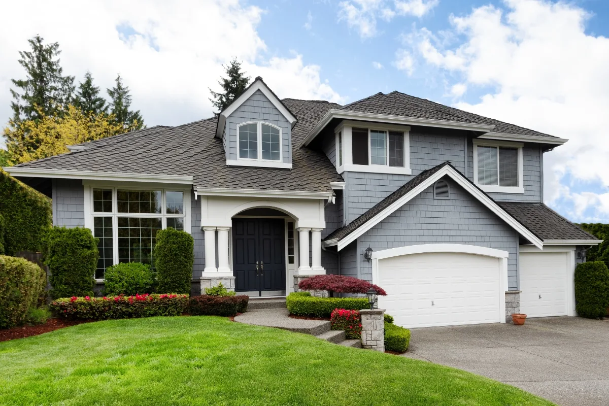 A two-story house with gray siding, white trim, and a double garage. The front yard makeover features lush green grass, stylish shrubs, and a few small trees that enhance its curb appeal.