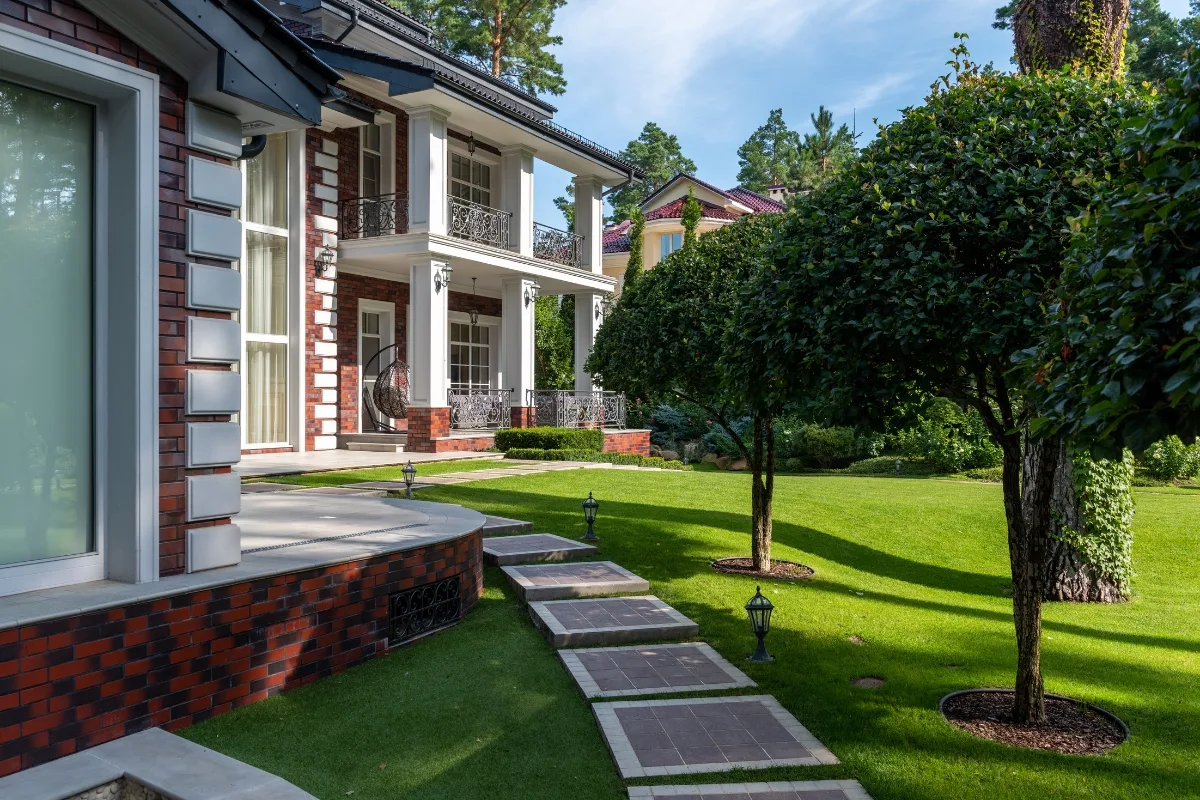 Front Yard Makeover: Transform Your Home With Low-Maintenance Landscaping Ideas 5 A brick house with large windows, a balcony, and an adjacent walkway showcases its stunning front yard makeover on a neatly manicured lawn with trees.