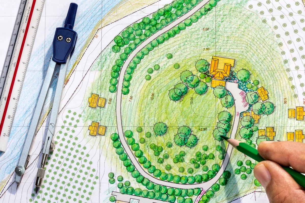 Front Yard Makeover: Transform Your Home With Low-Maintenance Landscaping Ideas 2 Hand drawing on a landscape design blueprint for a front yard makeover, complete with trees, pathways, and buildings. A compass, pencil, and ruler are nearby.