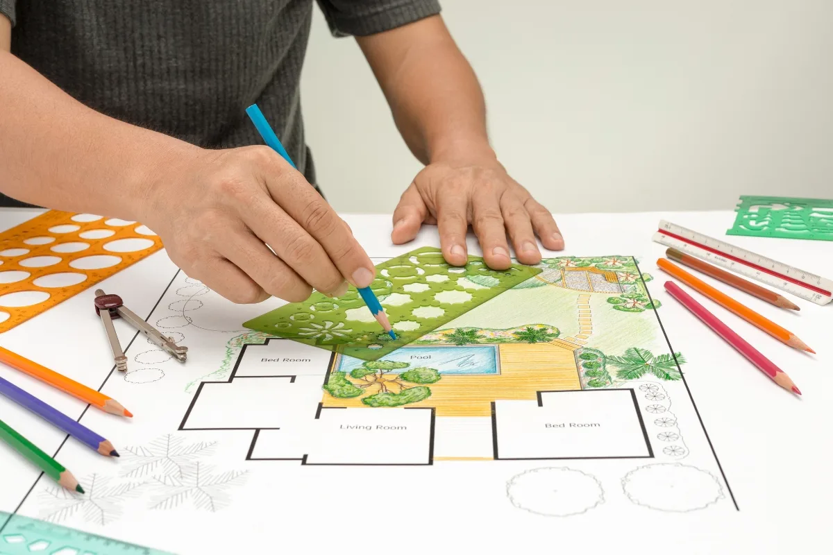A person is creatively drafting a landscape design with colored pencils and stencils on a table filled with drawing tools and geometric templates, subtly incorporating ideas on how to lay pavers into the vibrant plan.