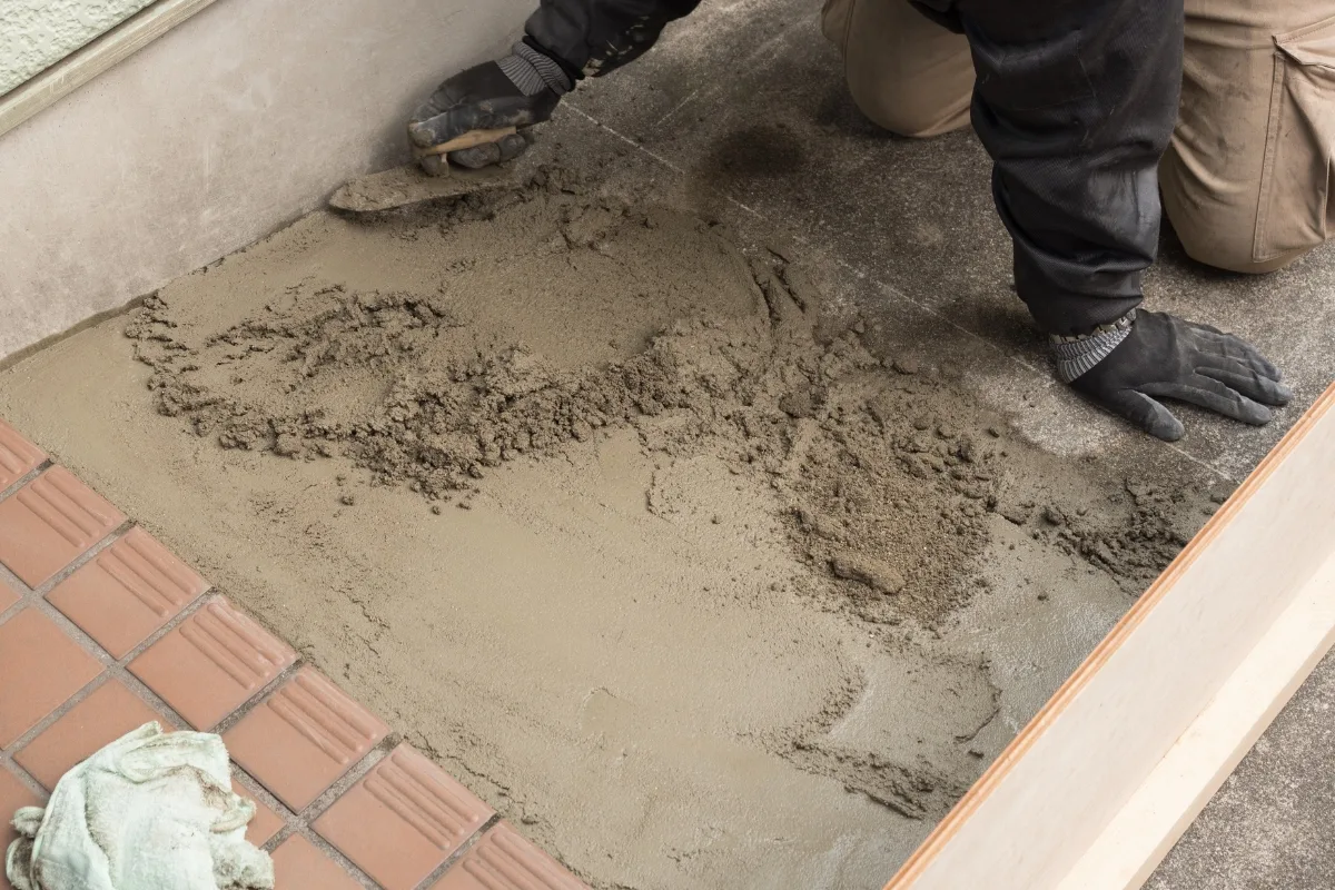 Kneeling, a person carefully smooths concrete with a trowel near the tiled area, laying the groundwork for knowing how to lay pavers. Dressed in work clothes and gloves, they ensure a perfect finish.