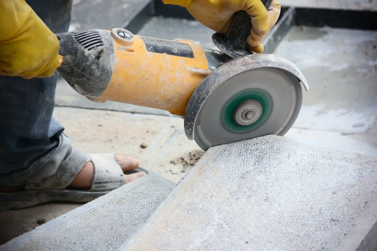 A person, prepped on how to lay pavers efficiently, uses a handheld electric grinder to cut through a concrete slab. Dust swirls around the tool as they skillfully prepare the surface for the forthcoming paving project.