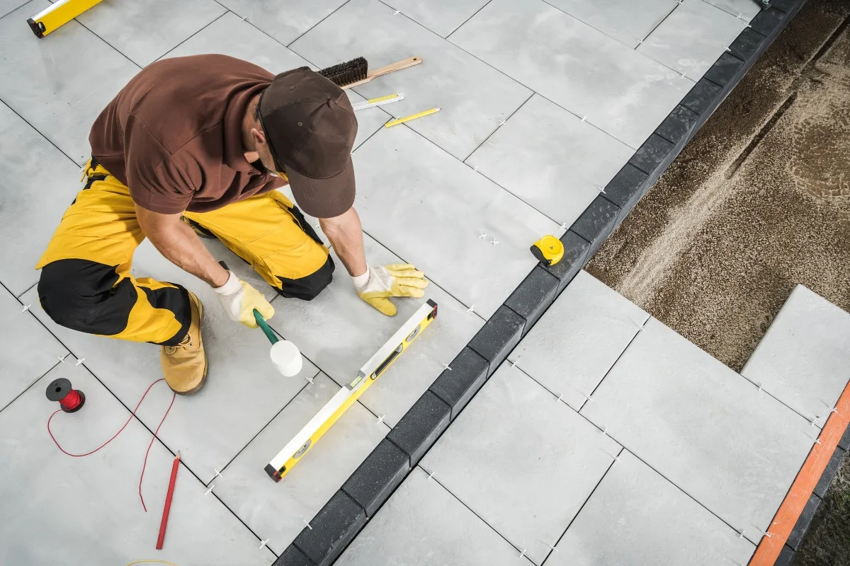 A worker in a brown shirt and yellow pants demonstrates how to lay pavers, using a mallet and level to install large square tiles on an outdoor surface. Tools and measuring tape are visible nearby.