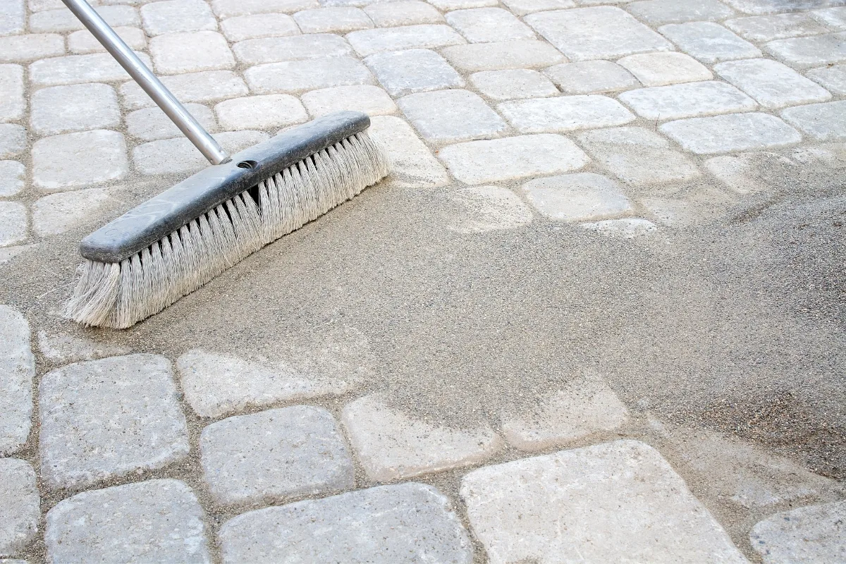 A broom briskly sweeps sand over stone pavers, echoing the final step in mastering how to lay pavers perfectly.