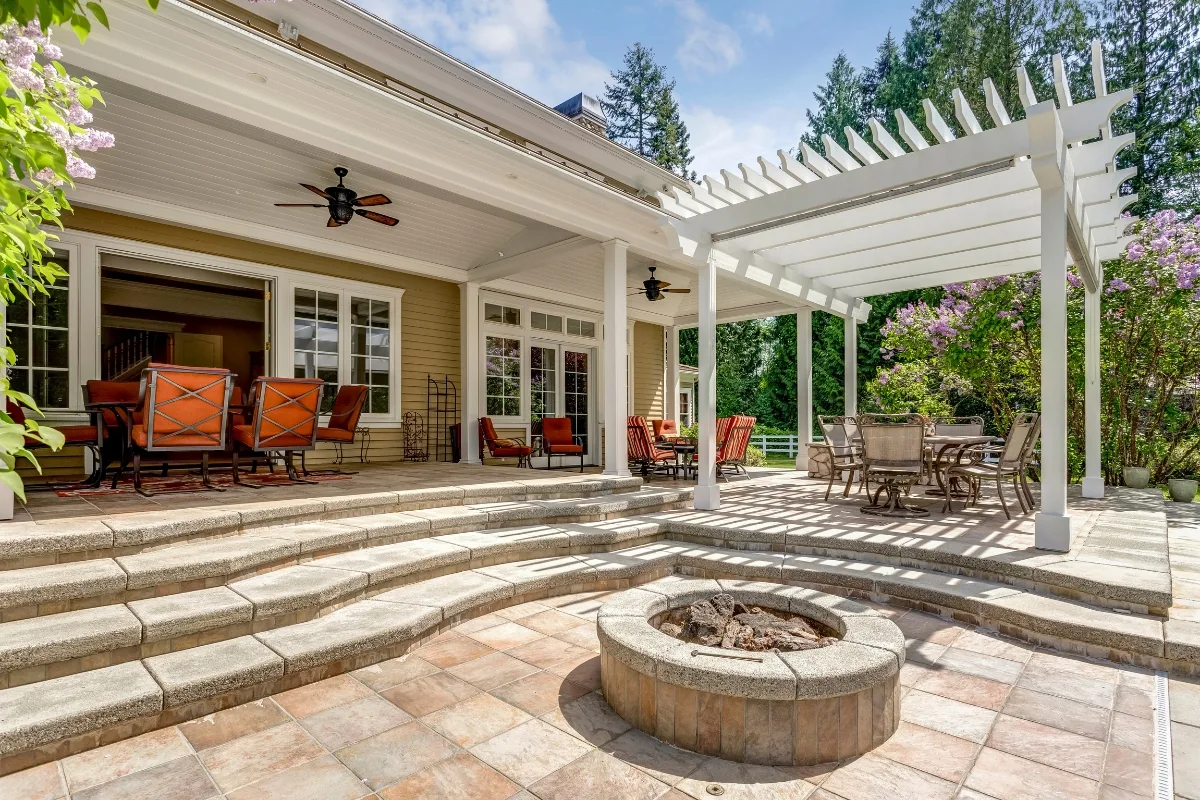 The backyard patio boasts a pergola, dining set, cushioned chairs, and a circular fire pit. Surrounded by trees and flowering shrubs under a clear sky, it's the perfect retreat. Discover how to lay pavers to enhance your own outdoor haven with these inviting features.