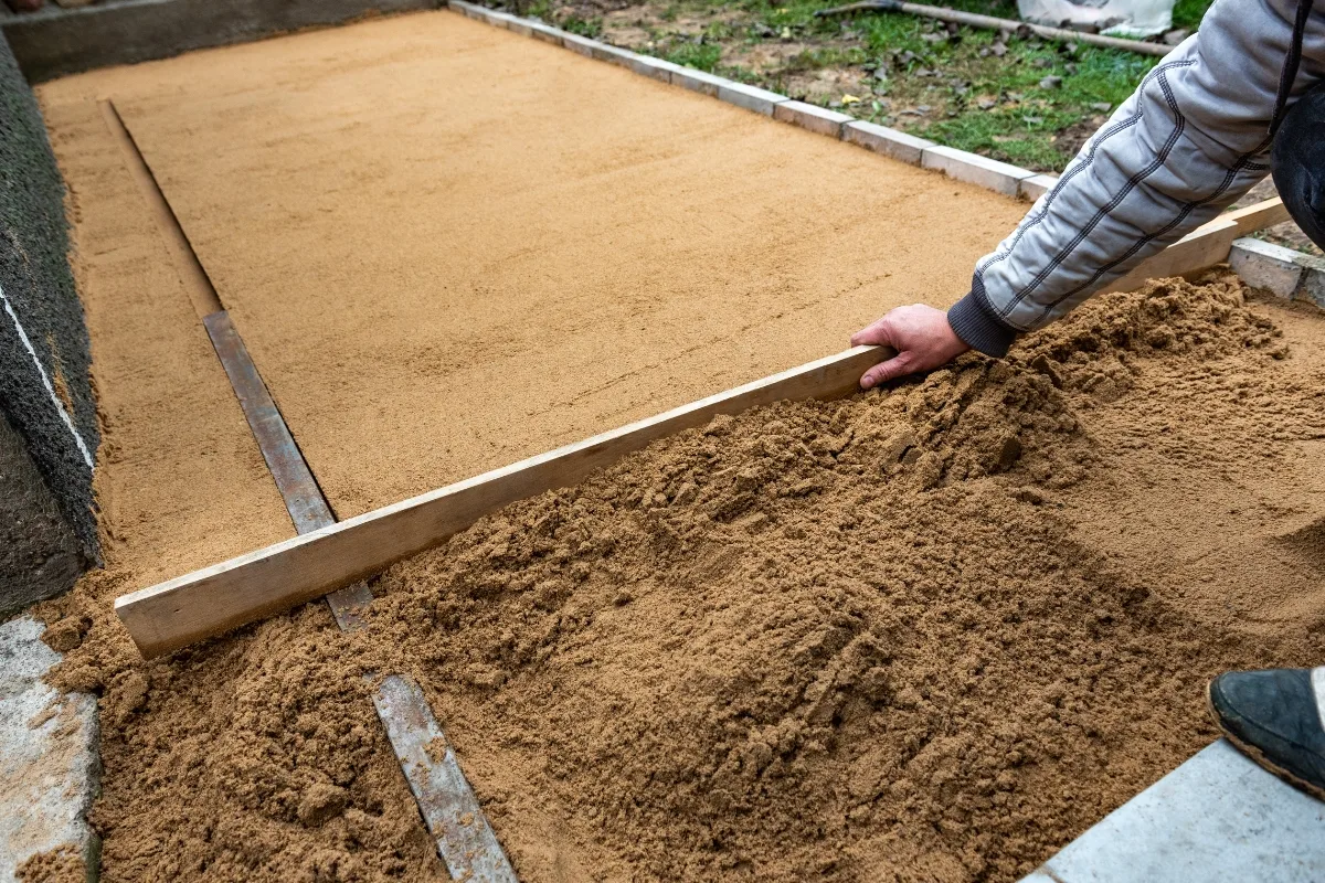 A person demonstrates how to lay pavers by meticulously leveling a layer of sand with a wooden board in a rectangular area.