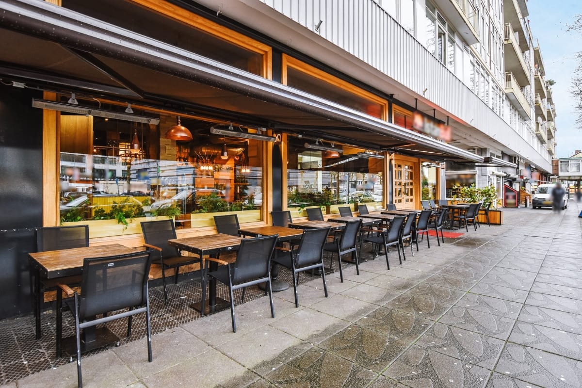 Outdoor Living Spaces in Woodstock: A Must for Local Businesses 4 Outdoor seating area of a modern restaurant featuring empty tables and chairs along a wet city sidewalk, large windows displaying stylish interior lighting and decor—perfect for enjoying vibrant outdoor living spaces.