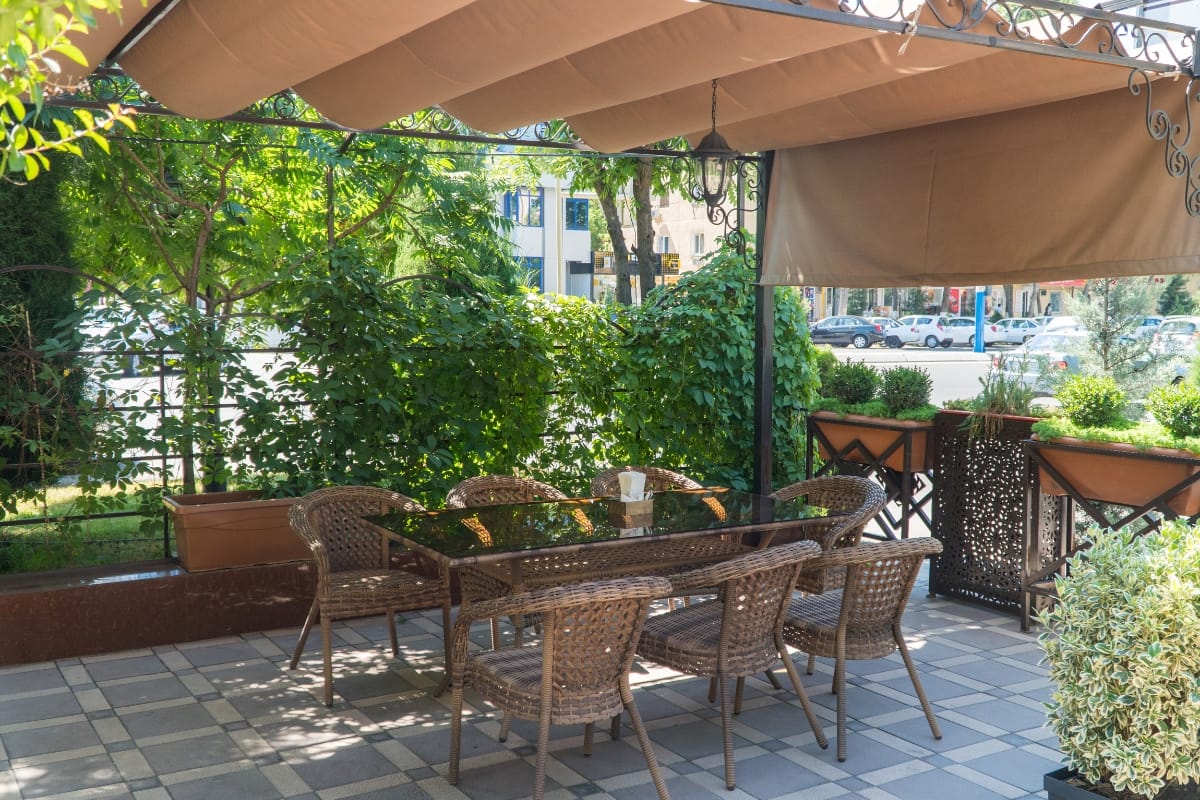 Outdoor Living Spaces in Woodstock: A Must for Local Businesses 3 Outdoor living spaces feature a glass-top table and six wicker chairs under a brown canopy, surrounded by lush green plants and planters on a tiled patio.