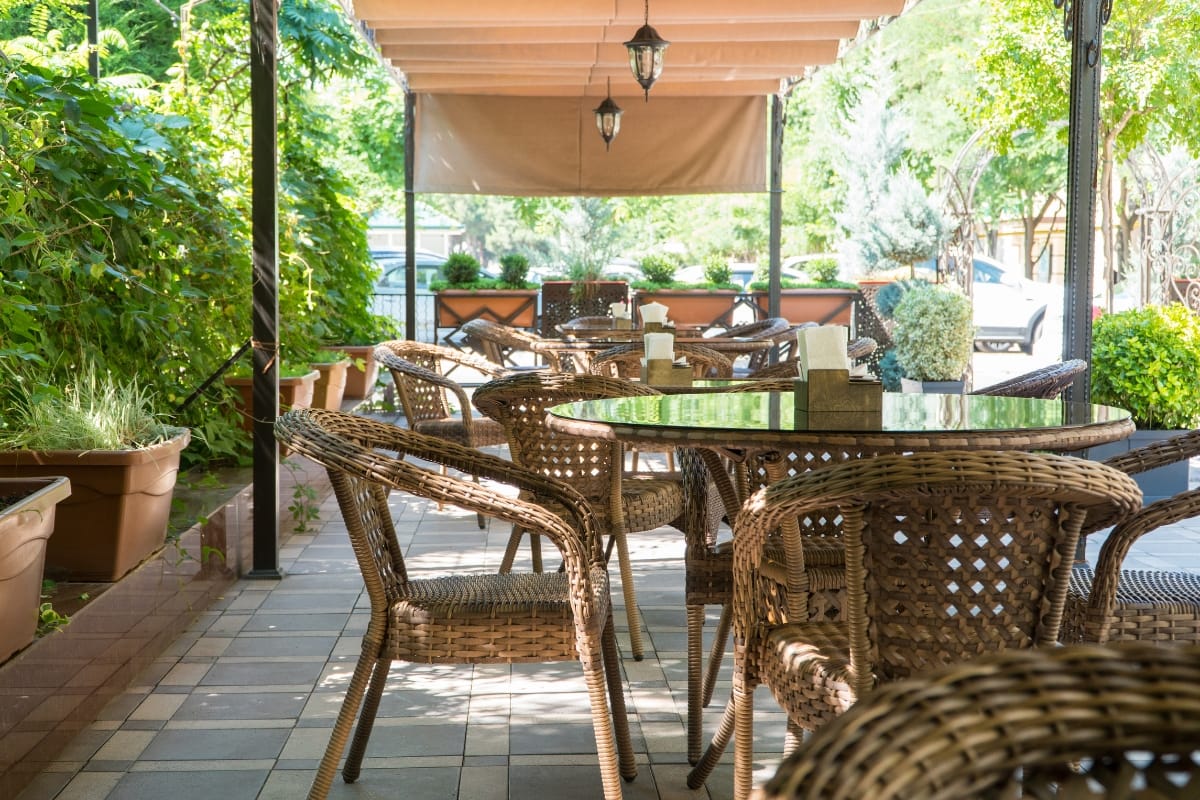 Outdoor Living Spaces in Woodstock: A Must for Local Businesses 2 Outdoor patio area with wicker chairs and glass tables under a canopy, surrounded by plants and natural light—perfect for enjoying stylish outdoor living spaces.