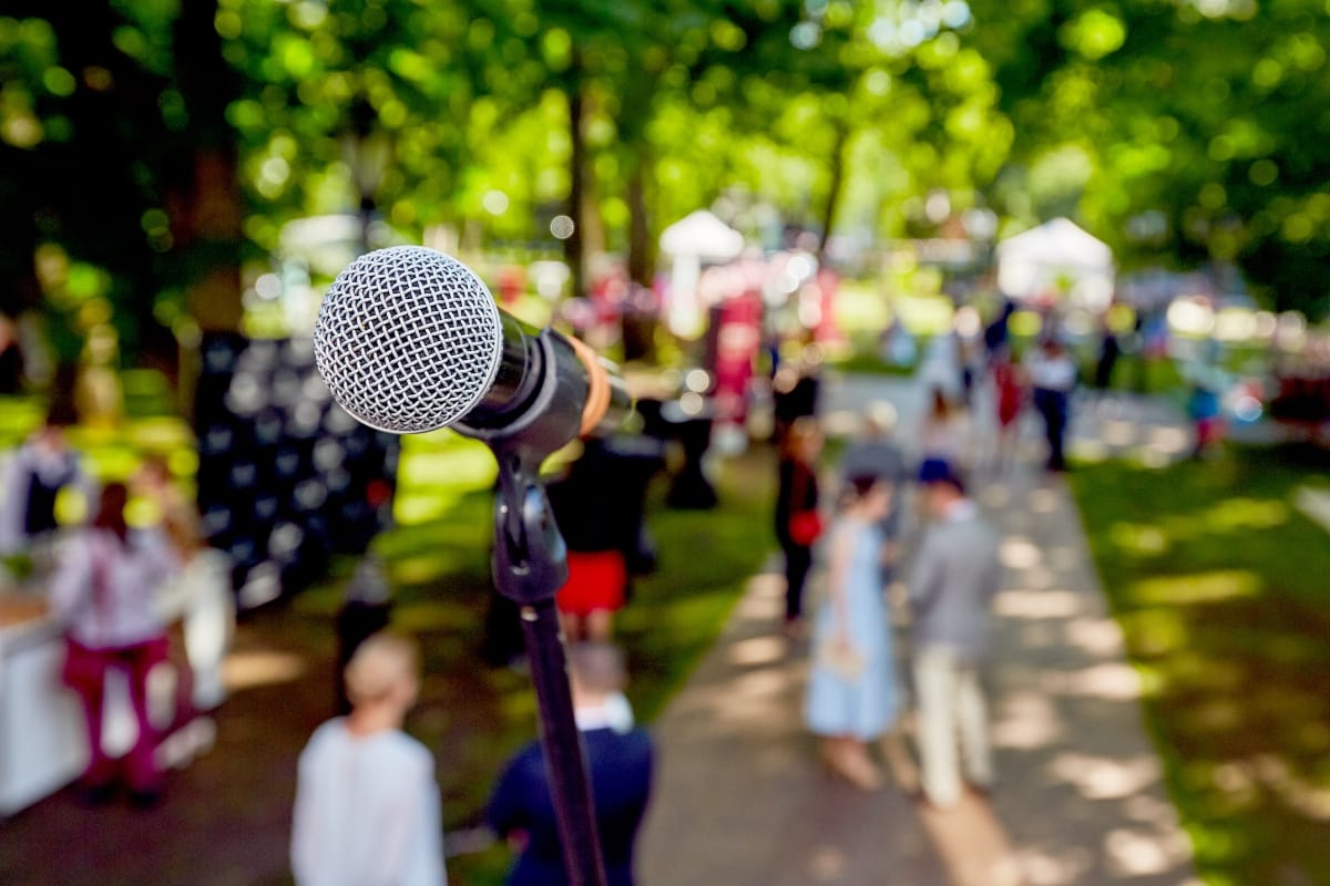 Outdoor Living Spaces in Woodstock: A Must for Local Businesses 4 Close-up of a microphone on a stand with people gathered in outdoor living spaces; trees and event tents create a lively park atmosphere in the blurred background.