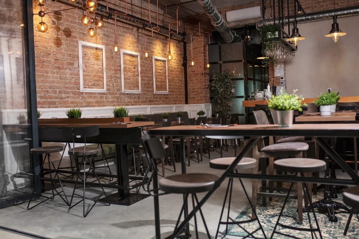Outdoor Living Spaces in Woodstock: A Must for Local Businesses 5 Modern cafe interior with exposed brick walls, empty tables and stools, hanging lights, potted plants, and an industrial-style ceiling—designed to bring the relaxed vibe of outdoor living spaces indoors.