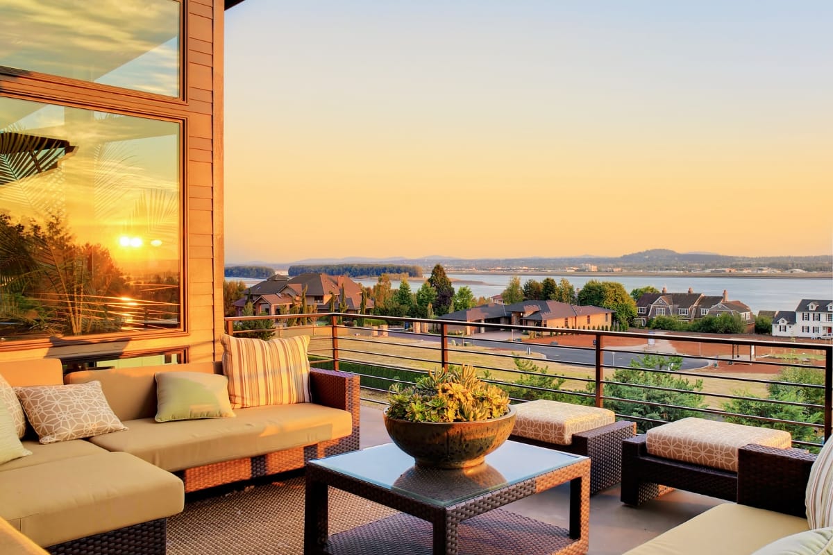 Outdoor Living Spaces in Woodstock: A Must for Local Businesses 7 Outdoor patio with cushioned seating and a large planter, overlooking houses, trees, and a body of water at sunset—perfect for relaxing in stylish outdoor living spaces.