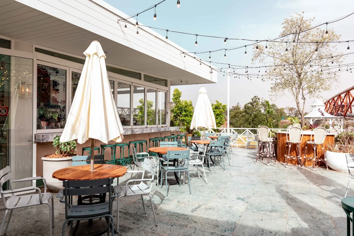 Outdoor Living Spaces in Woodstock: A Must for Local Businesses 1 Outdoor patio of a café with wooden tables, chairs, and white umbrellas. String lights hang above, creating inviting outdoor living spaces, with greenery in the background on a sunny day.