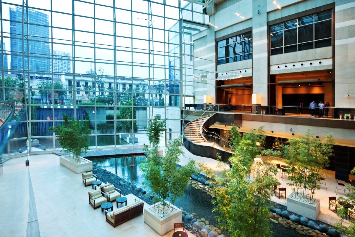 Commercial Water Features in Smyrna: How They Help Attract More Customers 4 Modern hotel lobby with large glass windows, commercial water features like an indoor stream, lush green plants, a seating area, and a curved staircase leading to an upper level.
