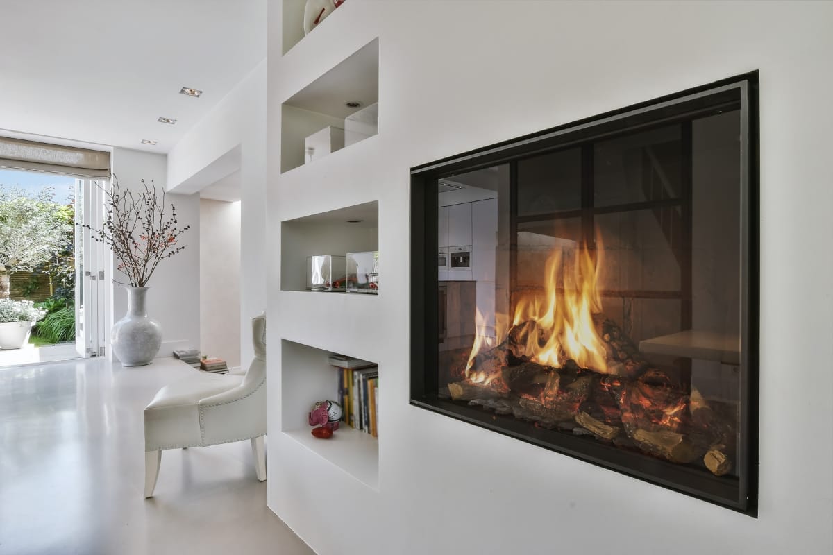 Fire Features in Buckhead: Why Commercial Spaces Should Invest 2 Modern living space with built-in shelves, a white armchair, large vase with branches, and a glass-enclosed fire feature set in a sleek white wall.