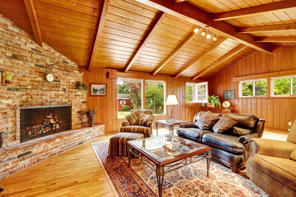 Fire Features in Buckhead: Why Commercial Spaces Should Invest 4 Cozy living room with wood-paneled walls, exposed beam ceiling, brick fireplace as a striking fire feature, patterned rug, armchair, leather sofa, glass coffee table, and large windows overlooking a green yard.