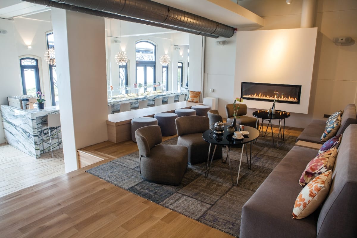 Fire Features in Buckhead: Why Commercial Spaces Should Invest 6 Modern living space with gray lounge chairs, a sectional sofa adorned with colorful pillows, a coffee table, striking fire features by the fireplace, and a dining area visible in the background.
