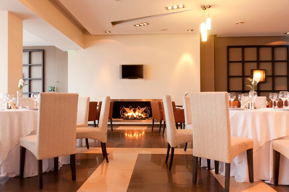 Fire Features in Buckhead: Why Commercial Spaces Should Invest 9 Modern restaurant dining area with beige chairs, round tables set with glassware and white tablecloths, and elegant fire features including a fireplace beneath a wall-mounted TV.