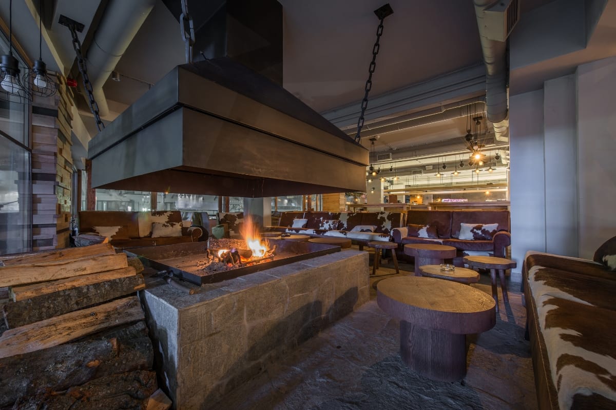 Fire Features in Buckhead: Why Commercial Spaces Should Invest 1 Modern indoor lounge with a central stone fireplace and other fire features, wooden tables, cushioned seating, and stacked firewood in a cozy, warmly lit setting.