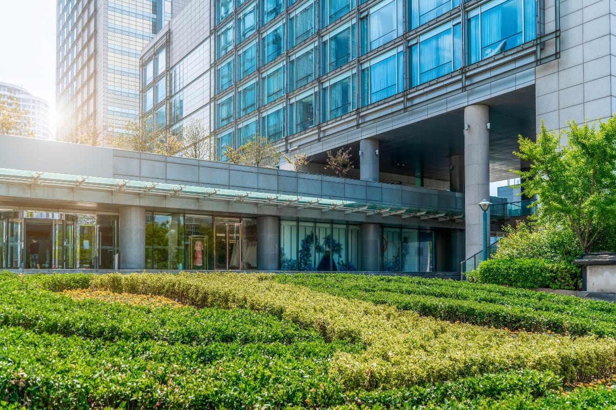 Commercial Landscape Maintenance for Alpharetta Businesses: Why It Matters 1 Modern office building with glass windows and a landscaped garden in the foreground, showcasing neatly trimmed green shrubs thanks to professional commercial landscape maintenance.