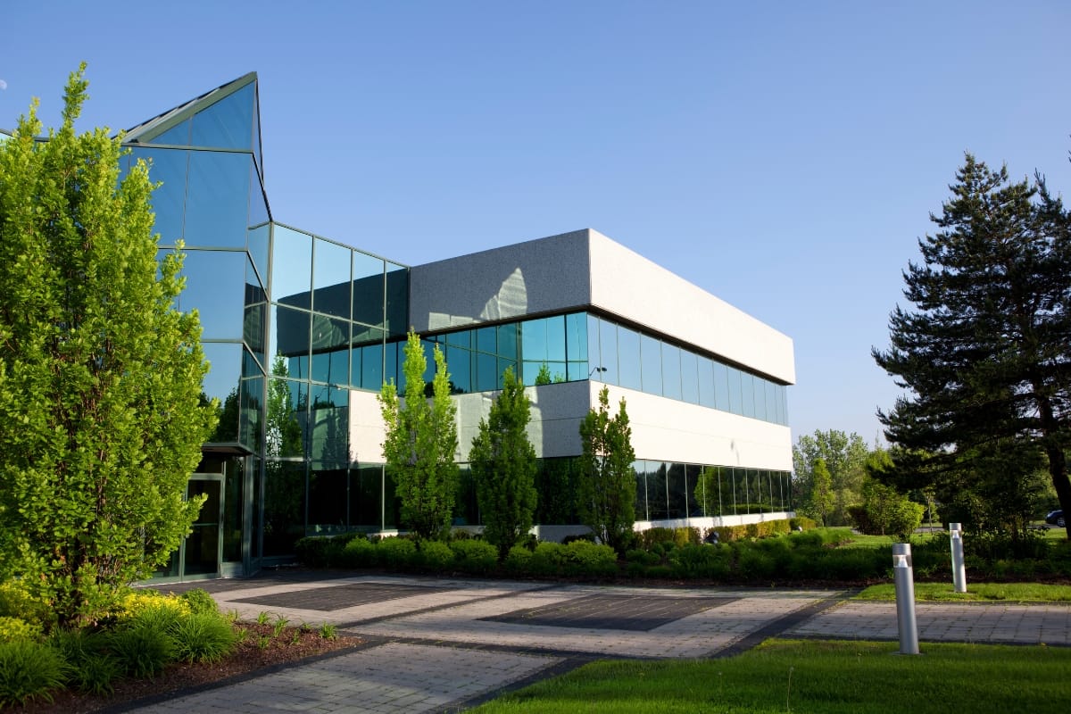 Commercial Landscape Maintenance for Alpharetta Businesses: Why It Matters 1 Modern office building with large glass windows, surrounded by trees and well-kept landscaping thanks to expert commercial landscape maintenance, under a clear blue sky.