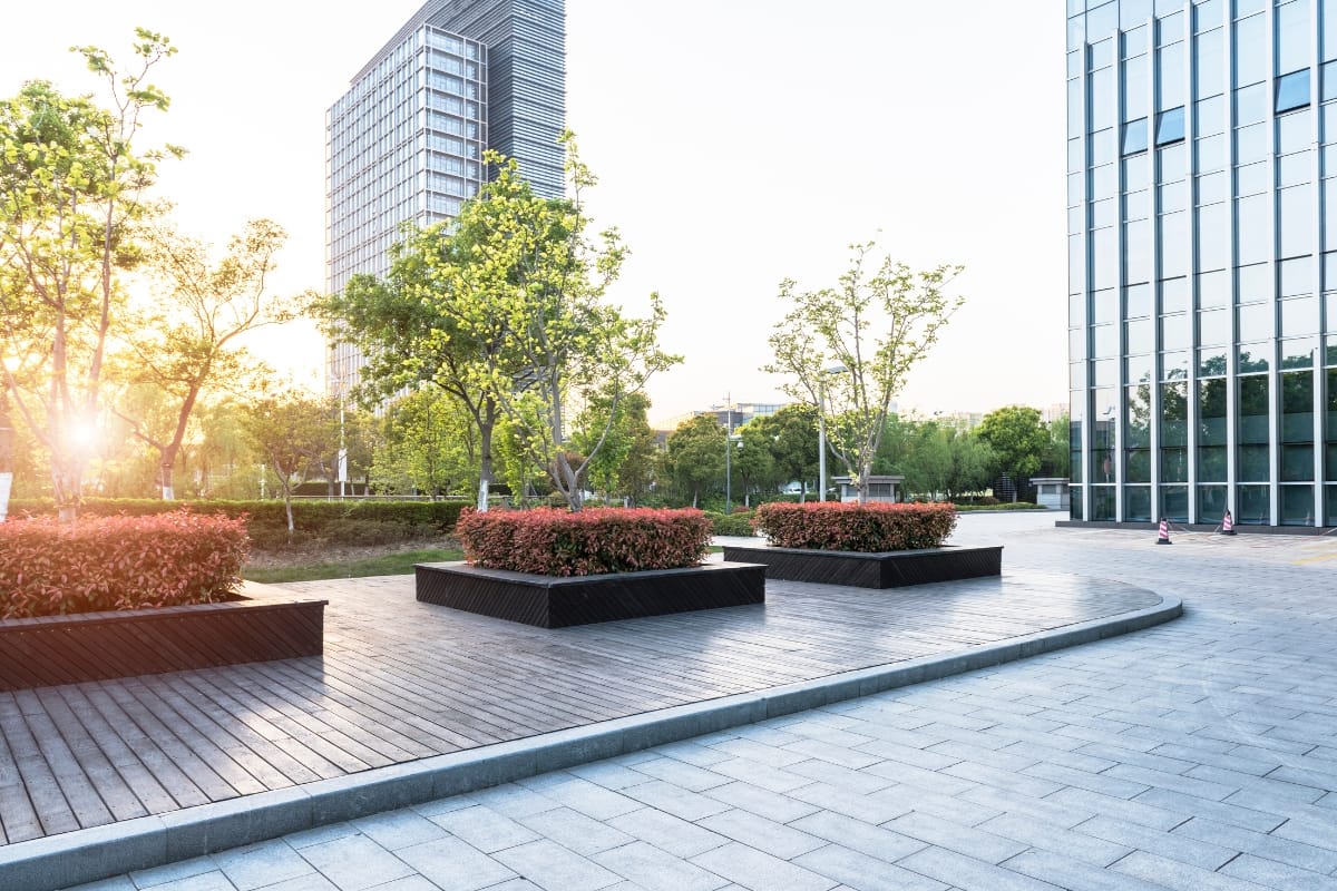 How Commercial Landscaping in Kennesaw Helps Create a Welcoming Business Environment 9 Modern outdoor plaza featuring commercial landscaping with wooden walkways, planter boxes filled with shrubs and trees, and tall glass office buildings at sunrise.