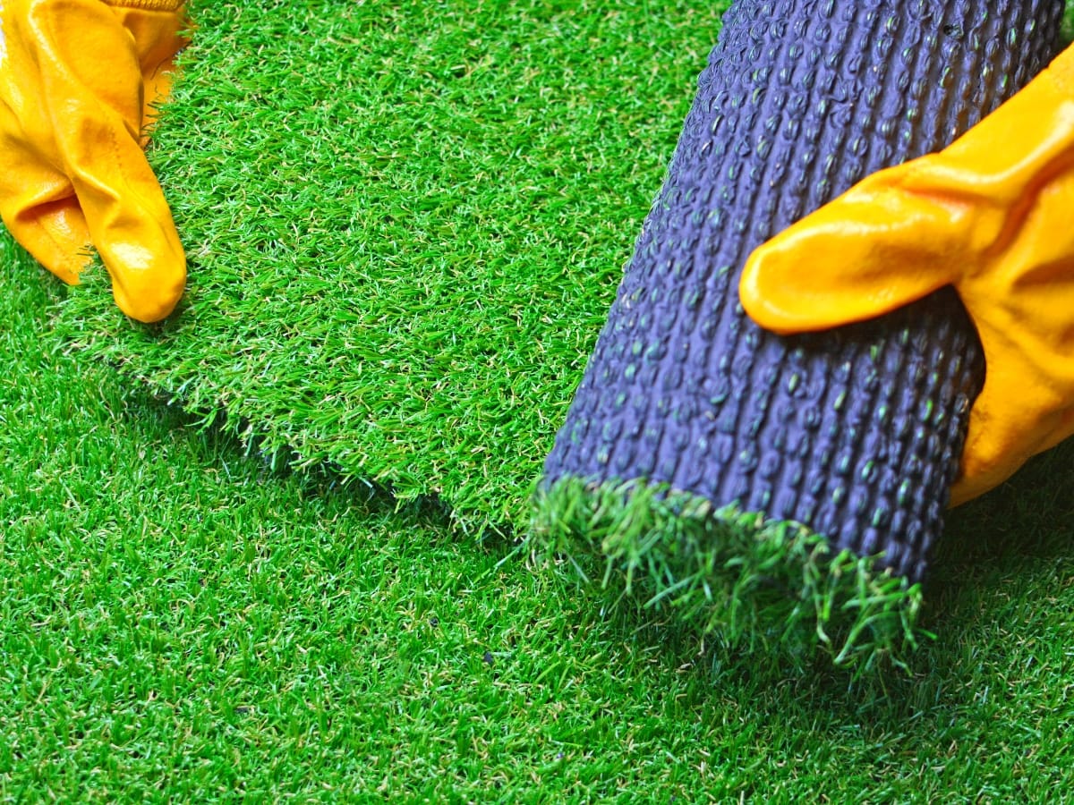 Hands in yellow gloves laying down a piece of artificial turf on top of a green surface.