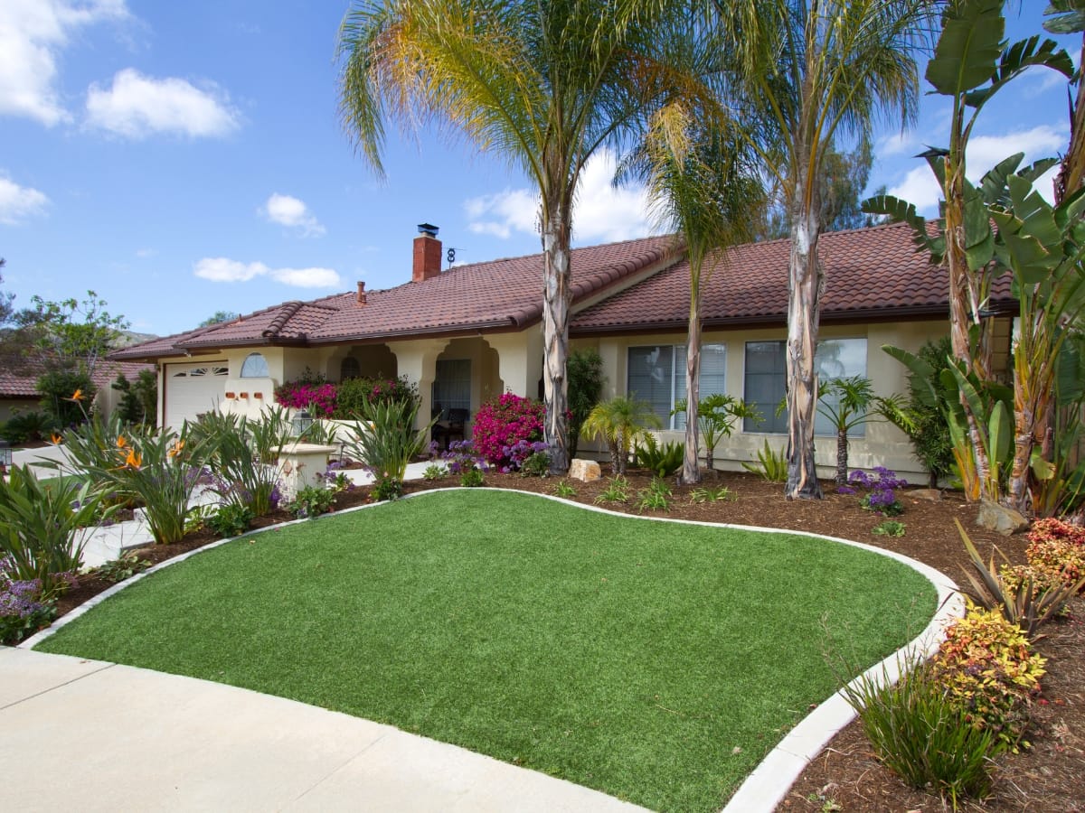 Single-story house with a red tile roof, palm trees, and front yard landscaping featuring a curved patch of artificial turf and a clear blue sky.