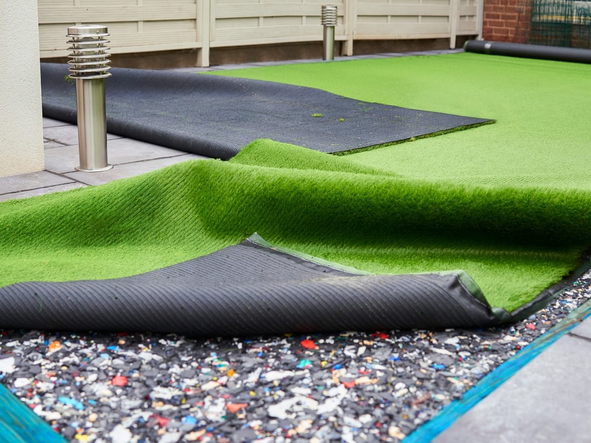 Rolled-up artificial turf with its backing exposed, laid on a hard surface with a visible recycled material layer underneath in an outdoor setting.