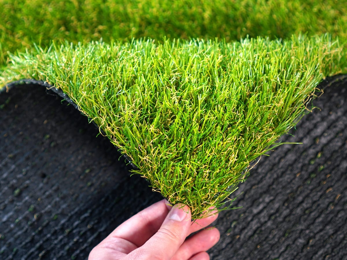 A hand lifts a corner of artificial turf, revealing vibrant green blades on top and a black textured backing underneath.